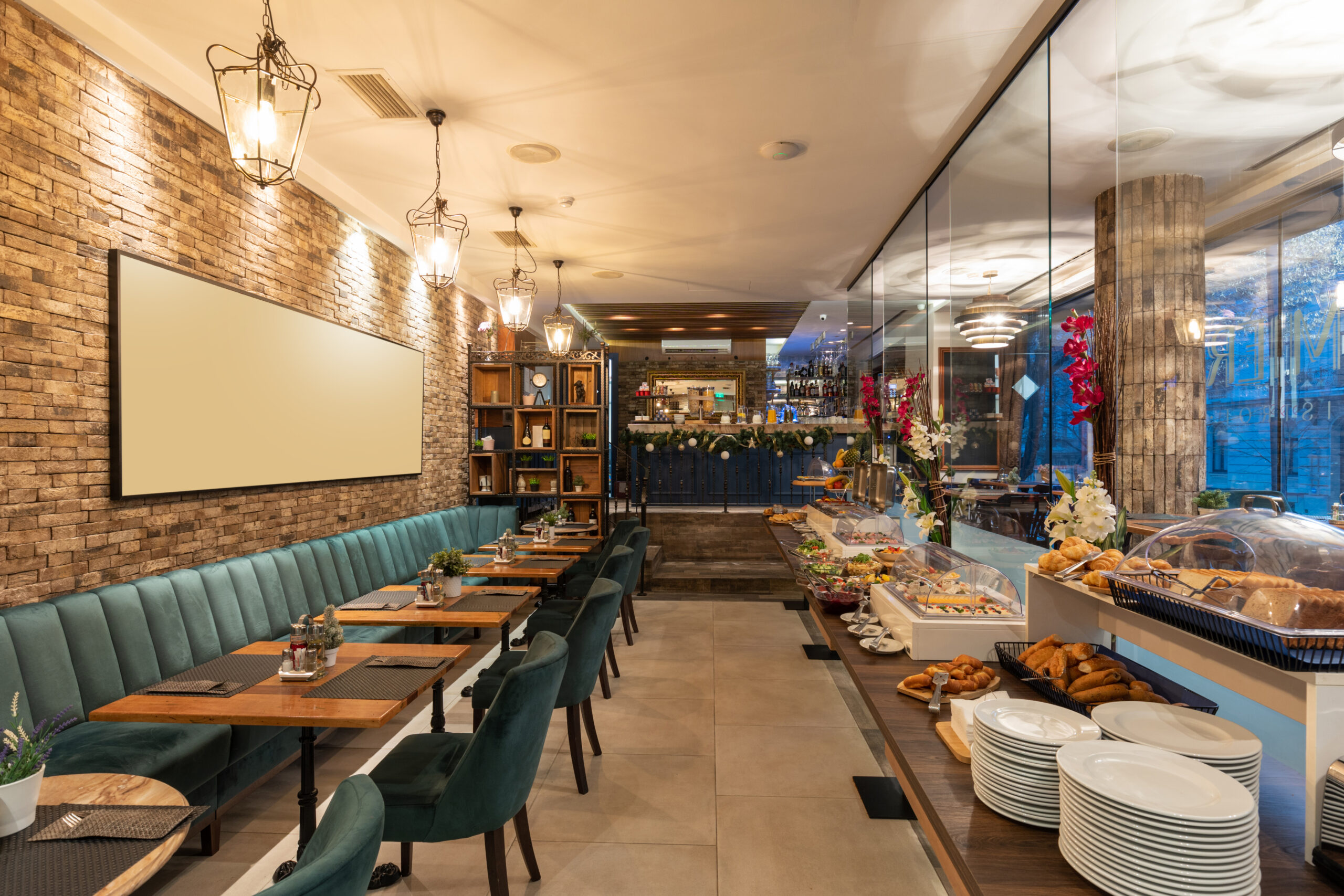 Interior de un restaurante elegante con decoración moderna. El restaurante cuenta con una pared de ladrillo a la vista, iluminación cálida, asientos acolchados en color verde esmeralda y mesas de madera. A la derecha, se observa una mesa buffet con una variedad de alimentos y platos dispuestos. Al fondo, se encuentra una barra decorada con flores y botellas.