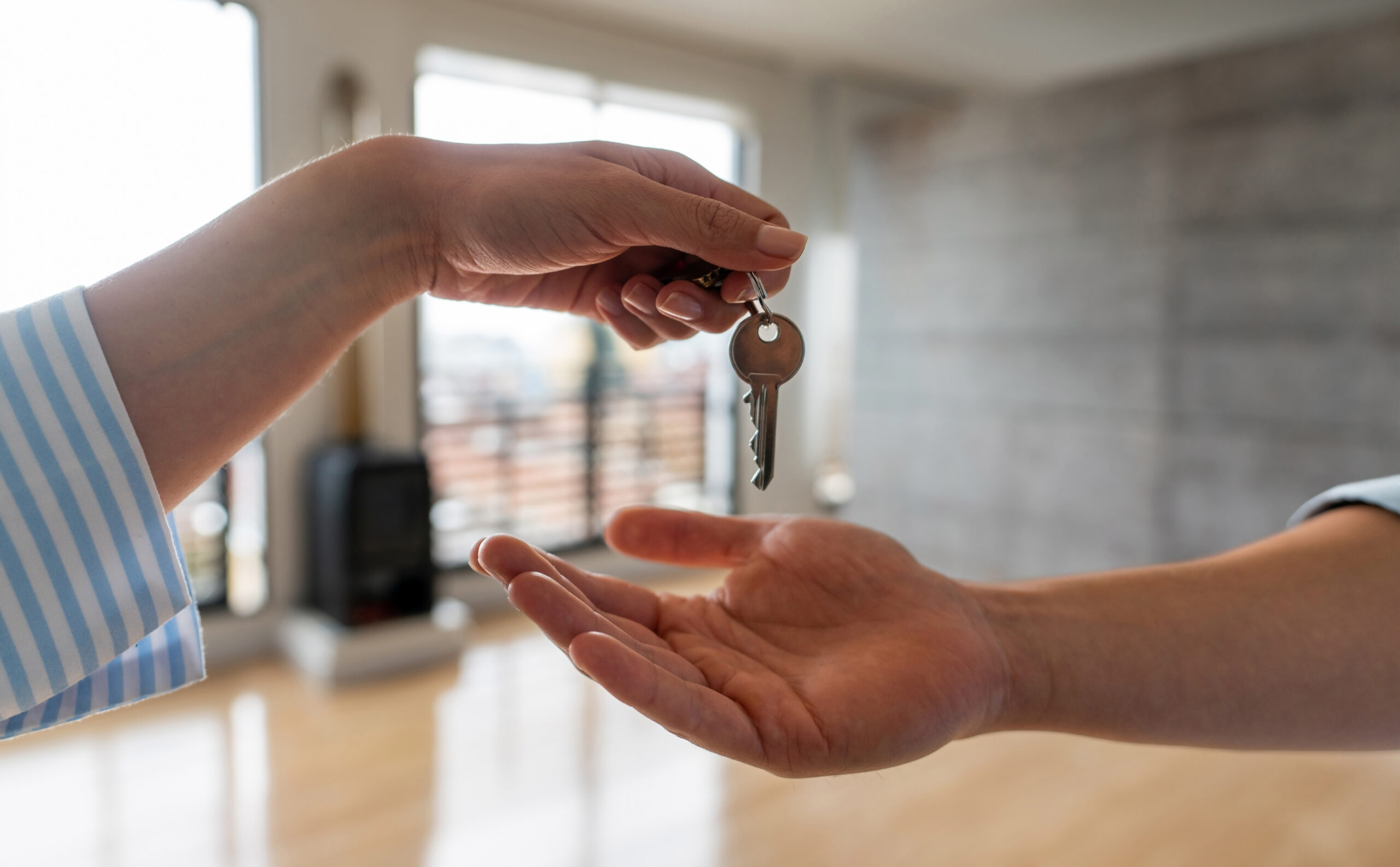 Un agente inmobiliario entrega las llaves de un nuevo piso al comprador. La imagen muestra un primer plano de las manos, con el fondo desenfocado de un interior moderno y luminoso. Esta escena representa el último paso en el proceso de compra de una propiedad, simbolizando la culminación de una transacción inmobiliaria exitosa.