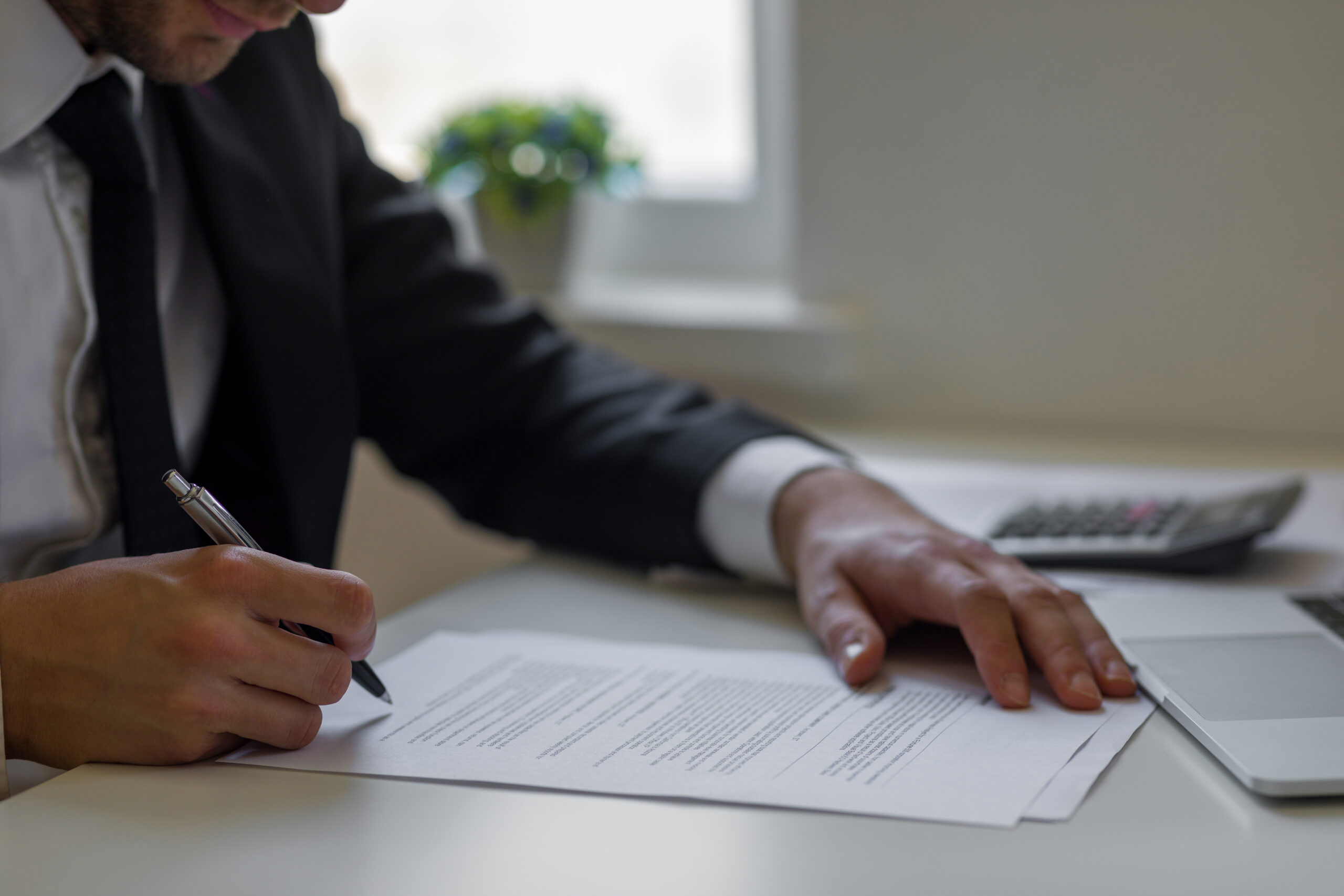 Hombre de negocios firmando documentos en una oficina. El hombre viste un traje oscuro y está concentrado en la firma de papeles sobre una mesa blanca. En la mesa hay una calculadora y una computadora portátil, lo que sugiere que está trabajando en asuntos financieros o legales.