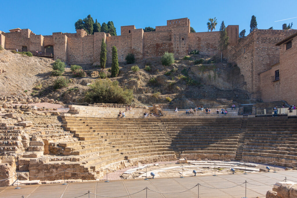 Vista del Teatro Romano de Málaga con la Alcazaba al fondo, una combinación única de arquitectura romana y fortaleza árabe, bajo un cielo despejado.