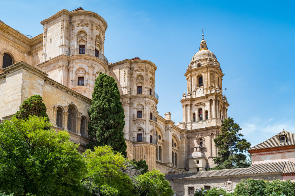 Primer plano de la Catedral de la Encarnación de Málaga, destacando su arquitectura renacentista y su emblemática torre, rodeada de vegetación y con un cielo despejado de fondo.