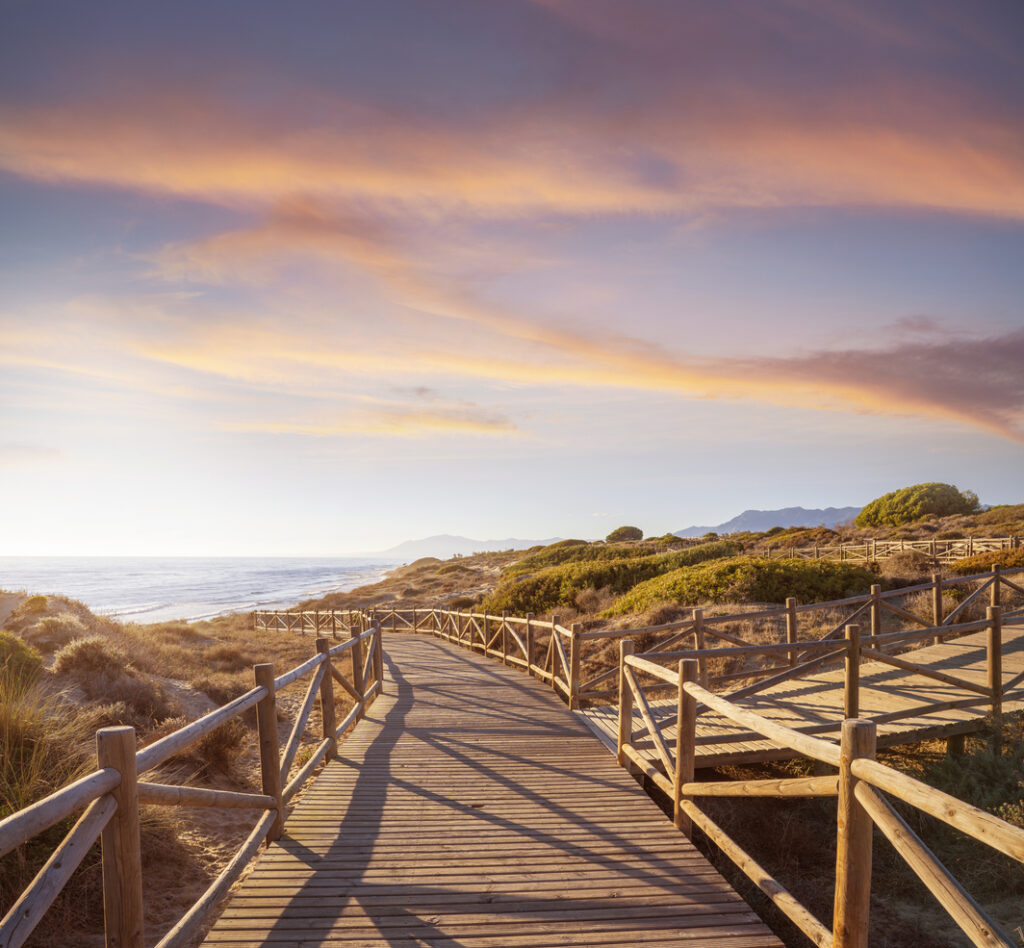 Pasarela de madera sobre las Dunas de Artola en Cabopino, Marbella, con vistas al mar Mediterráneo al atardecer y un cielo teñido de colores cálidos.