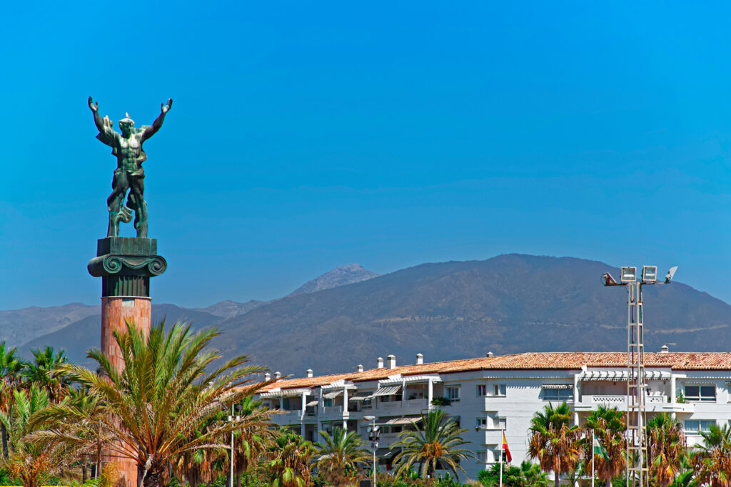 Estatua de la Victoria en Puerto Banús, rodeada de palmeras con las montañas al fondo bajo un cielo azul brillante.