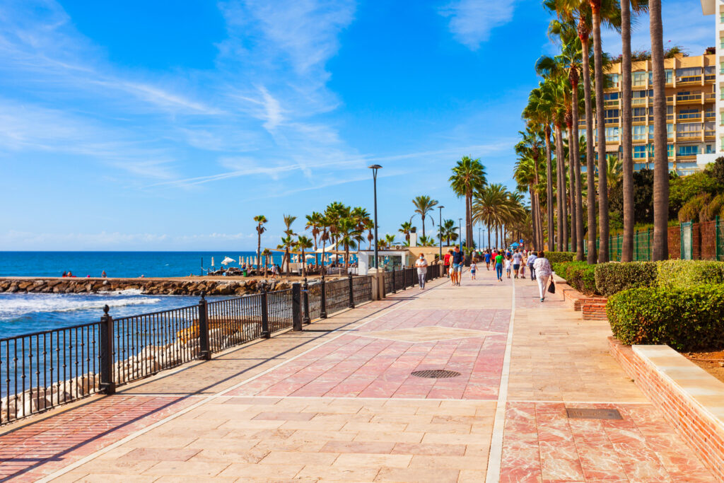 Paseo marítimo de Marbella con palmeras, vistas al mar Mediterráneo y personas disfrutando de un día soleado bajo un cielo azul.