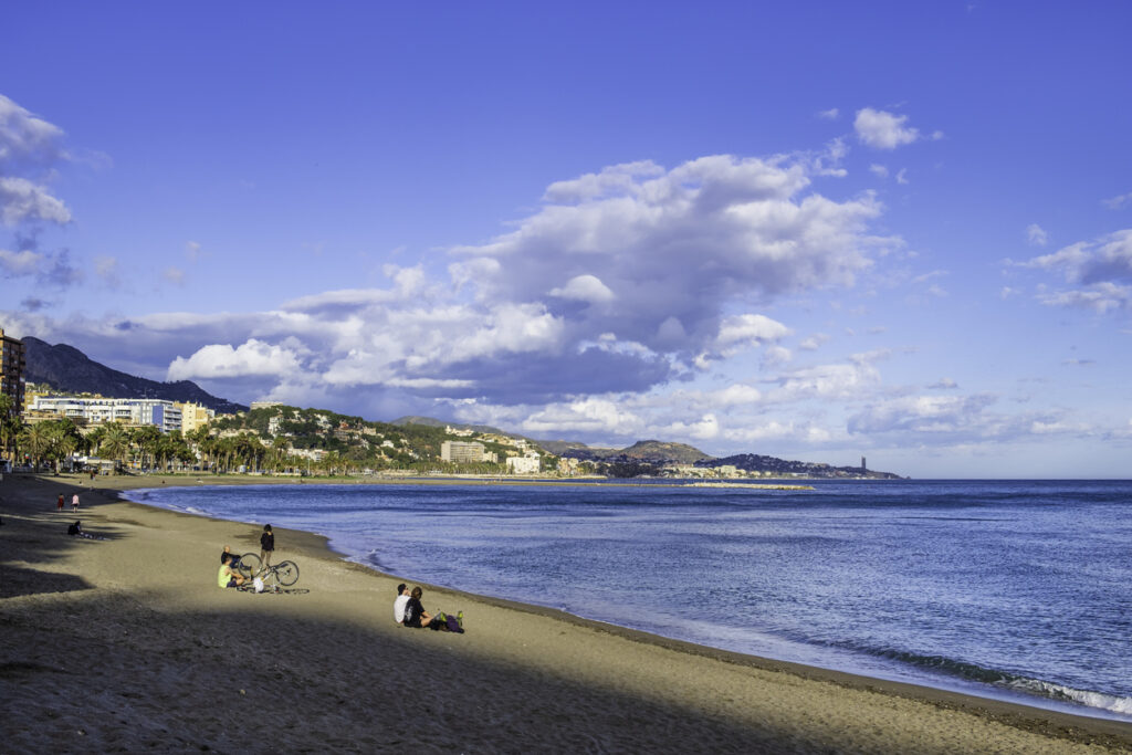 Vista panorámica de la Playa de La Malagueta en Málaga, con personas disfrutando del día junto al mar, palmeras y edificios al fondo, bajo un cielo azul con nubes esponjosas.