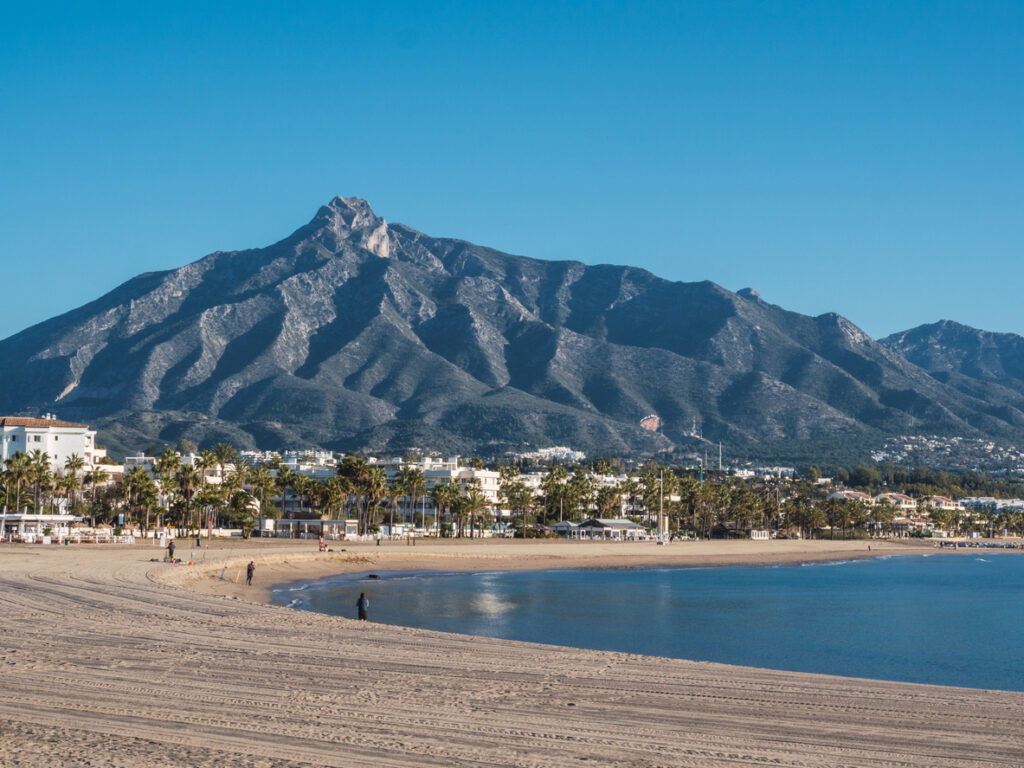 Playa de Puerto Banús con arena dorada, el mar tranquilo y la majestuosa Sierra Blanca al fondo bajo un cielo azul despejado