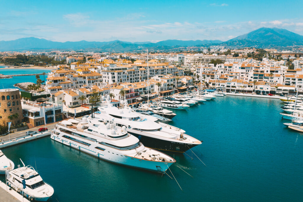 Vista aérea del puerto deportivo de Puerto Banús con yates de lujo, edificios blancos de estilo mediterráneo y las montañas al fondo bajo un cielo despejado.