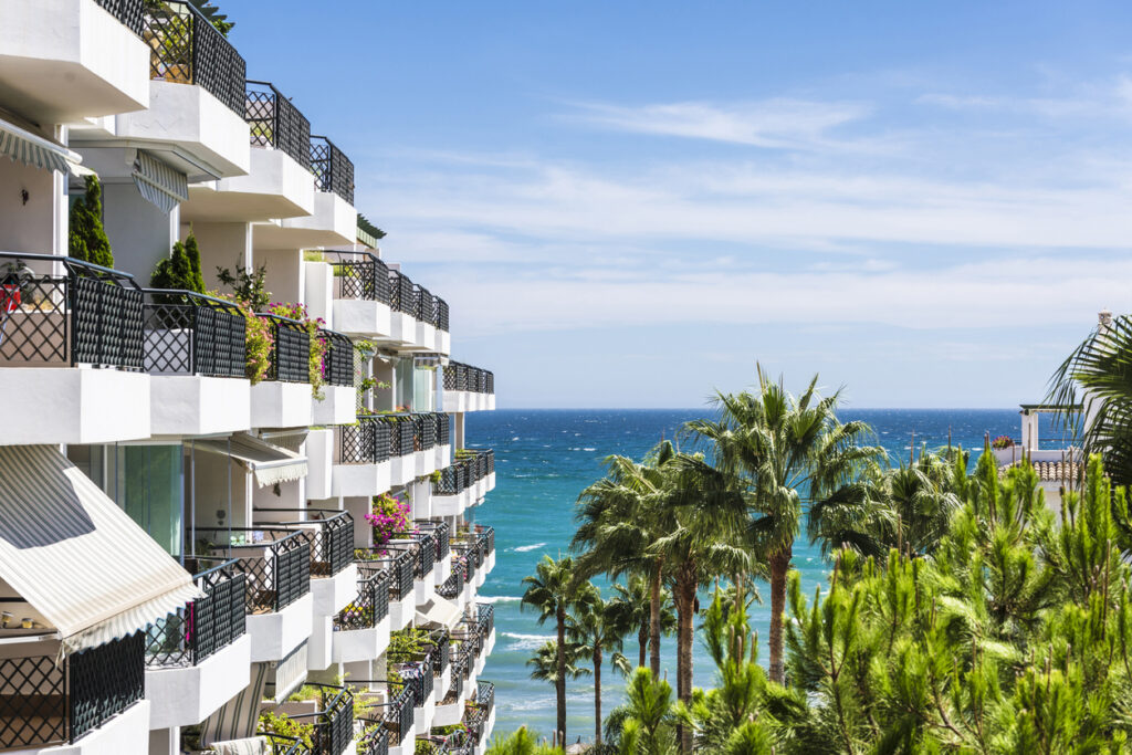 Fachada de apartamentos modernos en Málaga con balcones llenos de flores, rodeados de palmeras, con el mar Mediterráneo y un cielo azul de fondo.