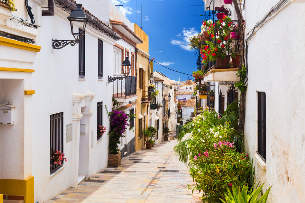 Callejón pintoresco en Marbella con fachadas blancas adornadas con flores coloridas y balcones tradicionales bajo un cielo azul.